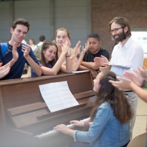 Teacher with group of students standing around piano and applauding